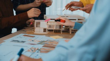 Çalışma alanındaki işbirliği ve yaratıcılığı vurgulayan, mimari bir model ve tasarım taslakları üzerinde çalışan elleri içeren bir ekip çalışması beyin fırtınası oturumunun dinamik bir sahnesi. SACTR