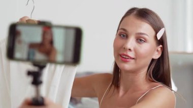 Genç sosyal medyum kadın moda videosu yapıyor. Blogcu kameraya selfie çubuğu ve ışık halkası kullanarak gülümserken, aynı zamanda ikna edici çevrimiçi kıyafetlerin izleyicilere veya takipçilere vlog satmasını sağlıyor. Düzenle