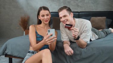 Genç çift, kredi kartıyla ödemek için internet üzerinden ödeme uygulaması ve dijital cüzdan kullanarak yatak odasında oturuyor. Mobil internet üzerinden alışveriş ve modern alımlar. Düzenle