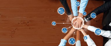 Çeşitli ellerin ahşap bir masanın üzerinde bir araya gelmesinin dinamik bir genel görünümü, çeşitli iş kavramlarını ve takım çalışmalarını temsil eden dijital simgelerle işbirliğini sembolize ediyor. Dostluk