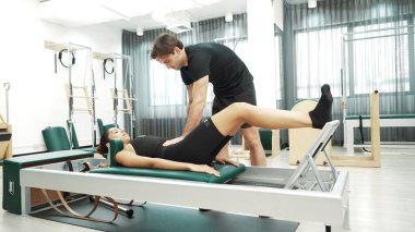 Mutlu pilates eğitmeni, bir müşteriye çekirdek istikrarı geliştirmek için bir reformcu üzerinde bacak egzersizi yapmasında yardımcı oluyor. Pilates makinesi kullanan sportif kız ark fıçısında kol dairesi pozu veriyor. Alışkanlık.