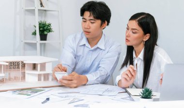 Profesyonel Asyalı erkek mimar ev modelinin uzunluğunu ölçmek için cetvel kullanırken, beyaz tenli genç meslektaşı ev modeliyle toplantı masasındaki verileri analiz etmek için dizüstü bilgisayar kullanıyor. Tertemiz..