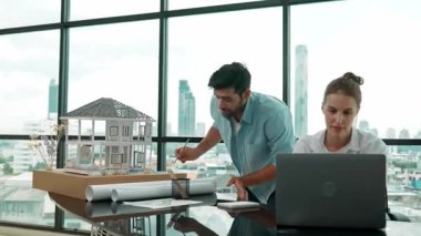 Akıllı mimar mühendis ev modelini incelerken meslektaşı da laptop analiz verilerini kullanıyor. Modern ofisteki ev modeli inşaatında profesyonel tasarımcı takımı birlikte çalışıyor. İzleme