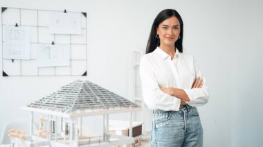 Genç, güzel, profesyonel mimar mühendisin portresini yakın plan ile birlikte beyaz modern ofiste ev modeliyle güven içinde pozlar veriyor. Yaratıcı yaşam ve tasarım. Tertemiz..