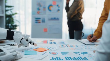 Gelecekçi bir robot eli modern bir iş toplantısında veri ve grafiklerle etkileşime girer, stratejik büyümeyi tartışan profesyoneller arasında işbirliği ve yenilik sergiler. Noogenesis
