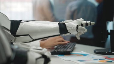 Modern ofis ortamında bilgisayar klavyesiyle etkileşime giren robotik bir kolun yakın çekimi, iş yerindeki teknoloji ve insan çalışmalarının karışımını vurguluyor. Noogenesis