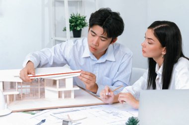 Profesyonel Asyalı erkek mimar ev modelinin uzunluğunu ölçmek için cetvel kullanırken, beyaz tenli genç meslektaşı ev modeliyle toplantı masasındaki verileri analiz etmek için dizüstü bilgisayar kullanıyor. Tertemiz..