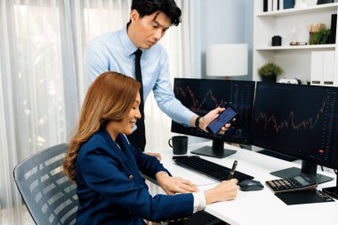 Gülümseyen işadamı cep telefonundaki dinamik borsa verilerini, iç ofisindeki internet sitesi uygulamasını analiz etmek için not yazan kadınlara gösteriyor. İnfobahn.