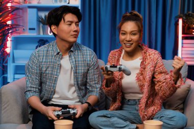 Mutlu çiftin oturduğu kanepe joysticklerle video oyunu oynuyor, meydan okuyan gülümseyen kadın, evdeki modern neon ışıklı oturma odasındaki mücadeleyi kazanmak için yumruk kaldırıyor. İnfobahn.