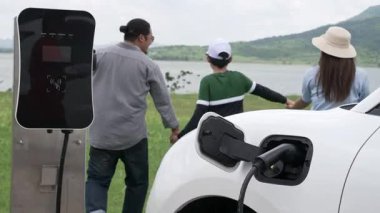 İlerici mutlu aile kavramı elektrikli araçla rüzgar çiftliğinde eğleniyor. Rüzgar türbini jeneratöründen gelen temiz yenilenebilir enerji ile şarj edilen elektrikli araç..