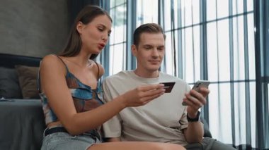 Genç çift, kredi kartıyla ödemek için internet üzerinden ödeme uygulaması ve dijital cüzdan kullanarak yatak odasında oturuyor. Mobil internet üzerinden alışveriş ve modern alımlar. Düzenle