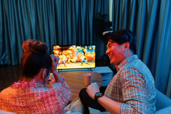 joystickli bir çift, TV ekranında dövüş oyunu oynuyor, erkek, kız arkadaşıyla yarışma seviyesini geri kazanıyor, modern evdeki neon ışıklı oturma odasında kanepeye oturuyor. İnfobahn.