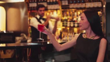 Güzel bir konuk, gece kulübünün mükemmel atmosferinde barmenlerin arka planında çiçeklerle süslenmiş karışık votka Cosmopolitan kırmızı kokteyli ile lüks bir gişede içki içerken kameraya bakıyor. Vinosity.