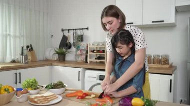 Zeki beyaz anne ve Asyalı kız birlikte yemek pişiriyor ve sebze doğruyor ya da akşam yemeği için salata hazırlıyor. Mutlu anne ve kız taze yemekle sağlıklı yemek yapıyorlar. Sağlıklı gıda konsepti. Pedagoji.