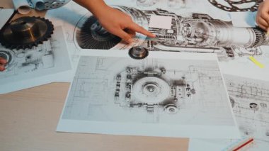 Modern bir atölyede, tasarım ve yenilikleri vurgulayan ahşap bir yüzey üzerinde makine mühendisliği planları ve viteslerle etkileşen ayrıntılı bir yakın çekim görüntüsü. SACTR