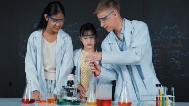 Zeki çocuk deney yaparken deney kabında sıvı karıştırıyor. Akademik öğrenci laboratuvar önlüğü giyerken masada mikroskop ve deneysel ekipmanlarla renkli örnekler koyuyor. Öğretim