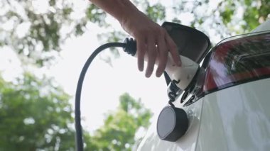 Yavaş çekim adamı, yaz mevsiminde açık yeşil yay yeşilliğindeki şarj istasyonundan elektrikli araba aküsünü şarj etmek için EV şarj aleti taktı. Yeniden şarj edilebilir elektrikli araç için sürdürülebilir enerji