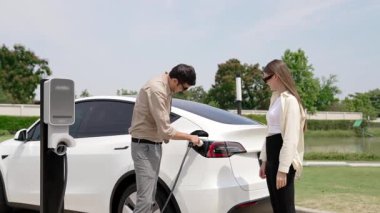 Yavaş çekim çift, ilkbaharda açık yeşil şehir parkındaki şarj istasyonundan elektrikli araba aküsünü şarj ediyor. Enerji sürdürülebilir kentsel seyahat yaşam tarzı için şarj edilebilir EV araba