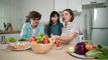 Çekici beyaz anne ve baba Asyalı kızıyla taze yemek pişirmek için birlikte vakit geçiriyorlar. Enerjik anne ve baba mutfakta salata yapmak için taze sebze hazırlıyor. Sağlıklı yiyecekler. Pedagoji.
