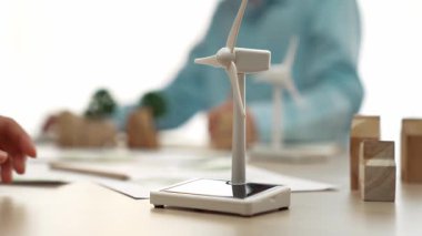İş kadını temiz enerjiyle temsil edilen yel değirmeni modelini tutuyor. Tahta blok sembolize ekoloji evi ve yeşil güç grafiği masanın üzerinde dağılıyor, iş adamı dizüstü bilgisayarla çalışıyor. Tanımlama.