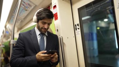 Akıllı yönetici trende dururken kulaklık takar ve müzik dinler. Profesyonel iş adamı, şarkıları dinlerken ve şarkı seçerken bölüme yaslanıyor. İş konsepti. Sevinçli..