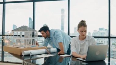 Akıllı mimar mühendis ev modelini incelerken meslektaşı da laptop analiz verilerini kullanıyor. Modern ofisteki ev modeli inşaatında profesyonel tasarımcı takımı birlikte çalışıyor. İzleme