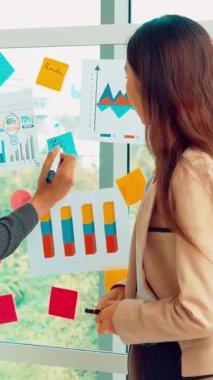 İş adamları ofisteki proje planlama kurulunda çalışıyor ve proje gelişimini analiz etmek için iş arkadaşlarıyla sohbet ediyorlar. Düzenli olması için cam duvara yapıştırılmış yapışkan notlar kullanıyorlar. .