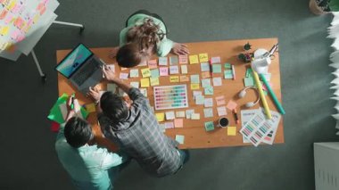 Gündelik kıyafet giyen, dizüstü bilgisayara bakan ve fikir paylaşan iş ekibinin zaman atlaması. Yapışkan notlar ve kulaklıkla masada çalışan ve renk paletini kontrol eden yöneticinin hava görüntüsü. Sempozyum.