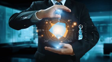 Takım elbiseli bir işadamı, yenilik ve dijital çözümlerin altını çizerek, modern ofis ortamında veri ve teknoloji elemanlarıyla çevrili 3 boyutlu bir holografik küp sergiliyor. Dörtgen