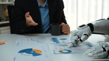 Bir insan ve robotik kol bir iş toplantısına katılır, masadaki veri tablolarını analiz eder, teknoloji ve insan zekası arasındaki işbirliğini etkili kararlar vermek için gösterir. Noogenesis