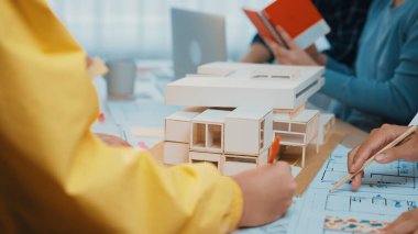 Bir grup profesyonel, tasarım planları ve modern ofis ortamında ayrıntılı bir model üzerinde çalışan bir mimari proje üzerinde işbirliği yapmaktadır. SACTR