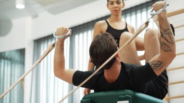 Müşteri, eğitmen desteğinin rehberliğinde kayışlarla Pilates reformatında egzersiz yapıyor. Sağlıklı bir adam pilates yapıyor ve üst vücut geliştirmek için Cadillac 'a asılıyor. Alışkanlık.