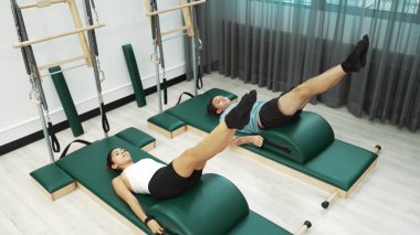 Pilates seansı sırasında iki müşteri yay namlu teçhizatı kullanarak bacak kaldırma pratiği yapıyor. Mutlu antrenör grubu karın kaslarını ve alt karın kaslarını geliştirmek için makas ya da bacak esneme pozu verir. Alışkanlık.