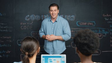 Tahtada çeşitli öğrencilere mühendislik kodları öğreten yakışıklı bir öğretmen. Mutlu çok kültürlü liseli çocuklar dizüstü bilgisayar kullanırken öğreniyor ya da programlama veya kodlama sistemini öğreniyor. Öğretim.
