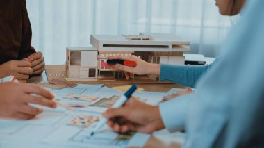 Bir ofis ortamındaki yaratıcılığı, takım çalışmasını ve tasarım sürecini vurgulayan, eskizler ve detaylı bir model üzerinde çalışan mimarların yer aldığı canlı bir işbirliği sahnesi. SACTR