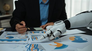 Çeşitli grafik ve grafikleri analiz eden bir robot kolu sergileyen fütüristik bir iş toplantısı. İş yerindeki teknoloji ve veri karışımının altını çiziyorum. Noogenesis