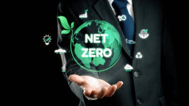 Bir işadamı, modern uygulamalar ve girişimlerdeki sürdürülebilirlik ve eko-arkadaşlığı gösteren yeşil simgelerle çevrili net sıfır hedefleri sembolize eden bir dünya sunuyor. Dörtgen