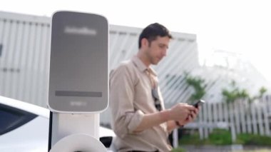 Genç adam, alışveriş merkezinin otoparkındaki EV otomobil şarj istasyonunda elektrik için akıllı telefon kullanıyor. EV aracıyla modern çevre ve sürdürülebilir kentsel yaşam tarzı