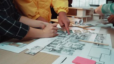 Stratejik proje toplantısı sırasında çeşitli bireylerin mimari planları ve modelleri analiz ettiği yoğun bir çalışma alanı. Parlak ortam işbirliğini teşvik eder. SACTR