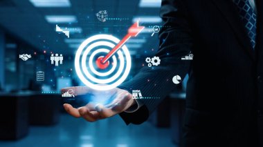Bir işadamı, yenilik ve büyüme fırsatlarını vurgulayarak, başarı ölçümleri ve stratejilerini temsil eden simgelerle çevrili çağdaş bir ofis ortamında dijital bir hedef gösteriyor. Dörtgen