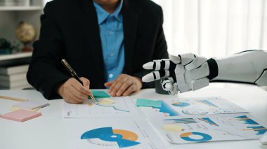 İnsan bir yazar, ofis ortamında çizelge ve verileri analiz eden robot bir el ile iş birliği yapar. Bu görüntü teknolojinin ve takım çalışmasının geleceğini temsil ediyor. Noogenesis