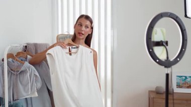 Genç sosyal medyum kadın moda videosu yapıyor. Blogcu kameraya selfie çubuğu ve ışık halkası kullanarak gülümserken, aynı zamanda ikna edici çevrimiçi kıyafetlerin izleyicilere veya takipçilere vlog satmasını sağlıyor. Düzenle