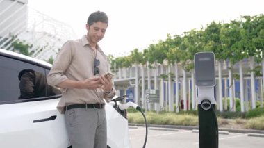 Genç adam, EV şarj aletini şehrin alışveriş merkezinin otoparkındaki şarj istasyonundan elektrik aküsünü şarj etmek için kullandı. Sürdürülebilir şehirde EV aracı tarafından şarj edilebilir enerji olarak kullanıldı.