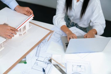 Profesyonel Asyalı erkek mimar ev modelinin uzunluğunu ölçmek için cetvel kullanırken, beyaz tenli genç meslektaşı ev modeliyle toplantı masasındaki verileri analiz etmek için dizüstü bilgisayar kullanıyor. Tertemiz..