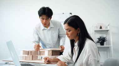 Profesyonel Asyalı erkek mimar ev modelinin uzunluğunu ölçmek için cetvel kullanırken, beyaz tenli genç meslektaşı ev modeliyle toplantı masasındaki verileri analiz etmek için dizüstü bilgisayar kullanıyor. Tertemiz..