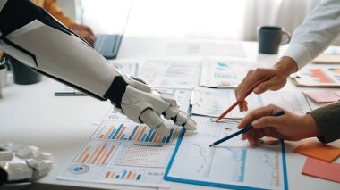 Bir robot kolu, modern ofis ortamında veri odaklı grafikler ve grafikler üzerinde, takım çalışmasını ve iş kararlarında teknolojik bütünleşmeyi sembolize eden bir insan kolu olarak uzanıyor. Noogenesis