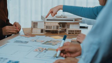 Bir grup mimar, parlak bir çalışma alanındaki bir masanın üzerinde bir ölçek modeli ve çoklu planların sergilenmesi hakkında yoğun bir tartışmaya girişiyor. SACTR