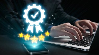 Bu resim, parlayan bir rozet ve yıldızlar aracılığıyla kalite güvencesinin dijital bir temsilini, dizüstü bilgisayarında yazan bir kişinin online başarıyı vurgulamasını içeriyor. Dörtgen