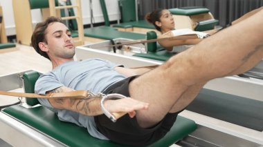 Bir grup müşteri, yenilikçi bir makinede bacak yükseltme işlemi yapıyor. Pilates partneri reformcu pozu veriyor ve egzersiz sırasında dengeyi geliştirmek ve koordinasyonu arttırmak için kayış takıyor. Alışkanlık.