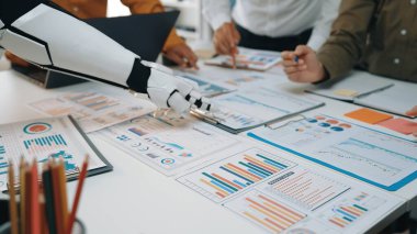 Robot bir kol ve insan elinin yer aldığı modern bir iş toplantısı sahnesi. Çeşitli çizelge ve raporları analiz etmek, takım çalışmasını ve teknoloji entegrasyonunu resmetmek. Noogenesis